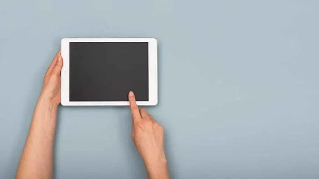 Une tablette blanche avec un écran noir est tenue et touchée par une main sur un fond bleu uni.