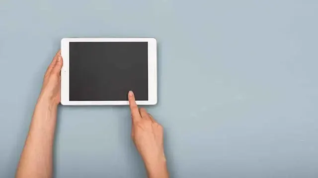 Une tablette blanche avec un écran noir est tenue et touchée par une main sur un fond bleu uni.