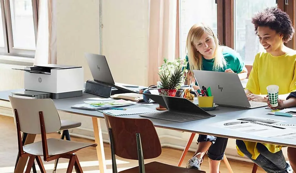 Twee laptops, een printer en kantoorbenodigdheden op een lange tafel in een lichte ruimte. Stoelen staan rondom.