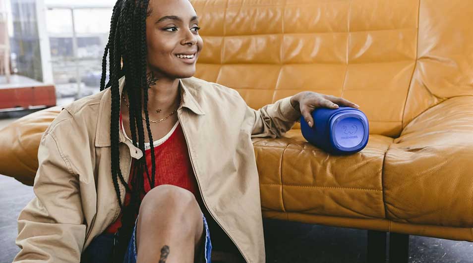 Une personne est assise sur un canapé jaune avec une enceinte Bluetooth JBL bleue à côté, portant une veste beige et un haut rouge.