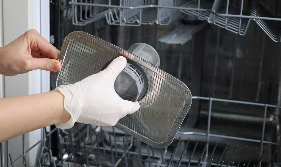 Gehandschoende handen halen een metalen filter uit een geopende vaatwasser. Binnenin zijn lege rekken zichtbaar terwijl het onderdeel wordt gereinigd.