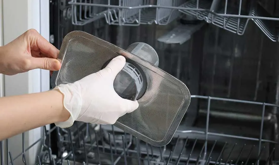 Gehandschoende handen halen een metalen filter uit een geopende vaatwasser. Binnenin zijn lege rekken zichtbaar terwijl het onderdeel wordt gereinigd.