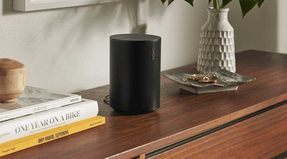 Zwarte Sonos speaker op houten kast, naast boeken, vaas en decoratieve schaal.