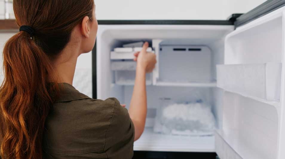 Une personne manipule un distributeur de glace dans un congélateur blanc ouvert, avec des étagères visibles.