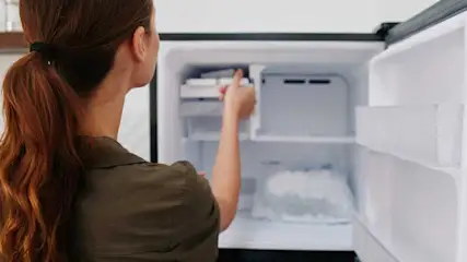 Une personne manipule un distributeur de glace dans un congélateur blanc ouvert, avec des étagères visibles.