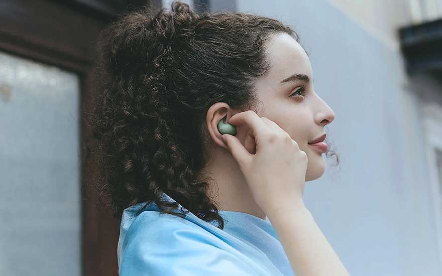 Young woman with curly hair inserts a green wireless earbud, listening to her favourite music while walking outside in the city.