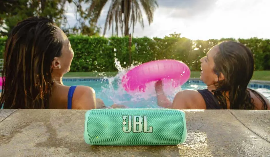 Sur le bord de la piscine se trouve une enceinte Bluetooth JBL vert menthe, pendant que deux personnes jouent dans l’eau avec une bouée rose.