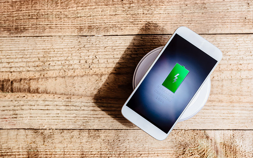 Een witte smartphone ligt op een witte draadloze oplader op een houten tafel, het scherm toont een groene batterij.