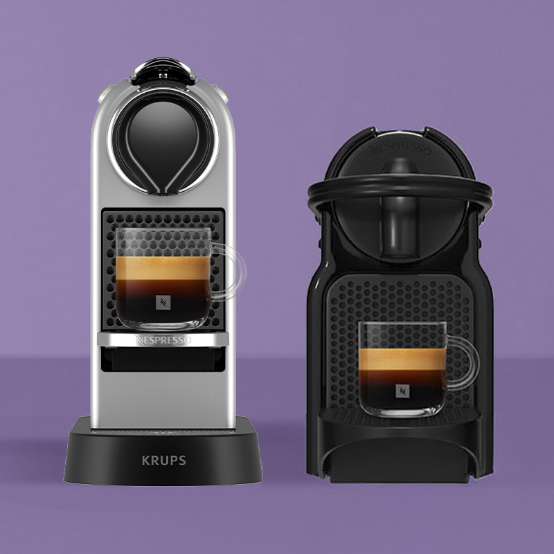 Een zilverkleurige en een zwarte Nespresso-koffiemachine met elk een espresso in een glazen kopje, op een paarse achtergrond.