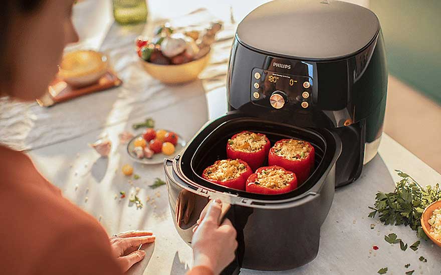 Une friteuse à air noire avec des poivrons rouges farcis est ouverte sur une table avec des ingrédients.