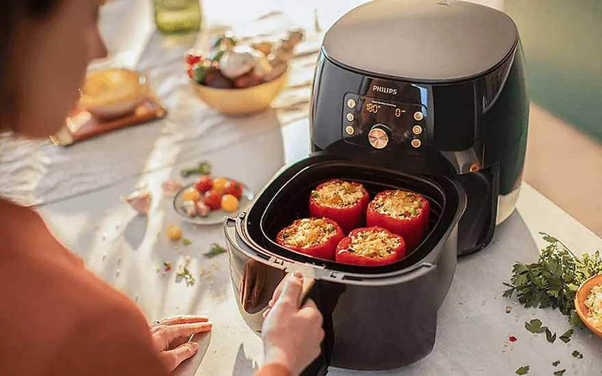 Une friteuse à air noire avec des poivrons rouges farcis est ouverte sur une table avec des ingrédients.