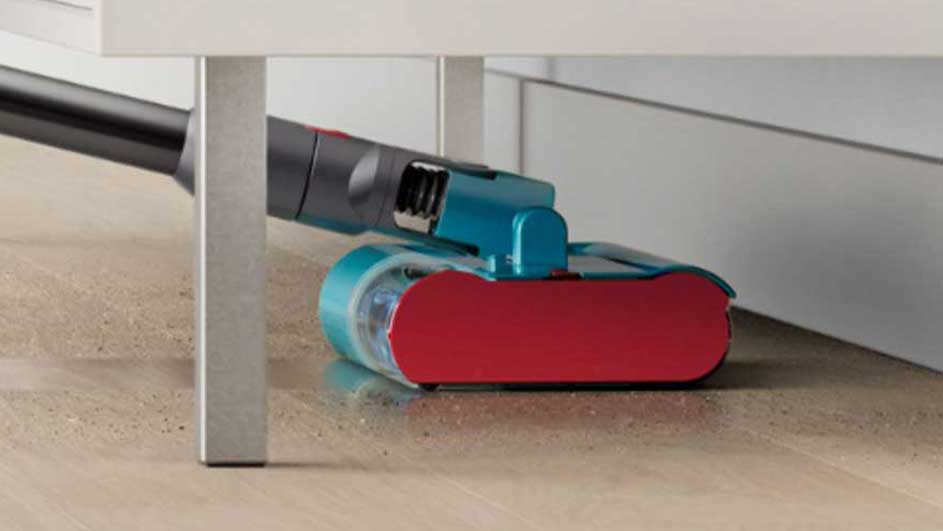 Un aspirateur rouge et bleu nettoie sous un meuble blanc. Le sol est beige clair.