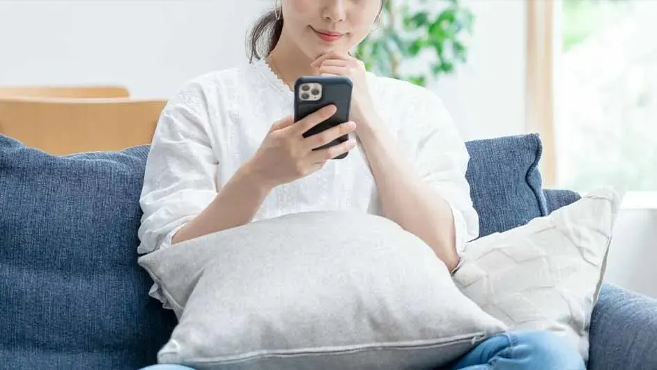 A woman sits on a sofa, leaning on cushions, calmly using an Apple iPhone with a dark case, soft daylight and houseplants in the background.
