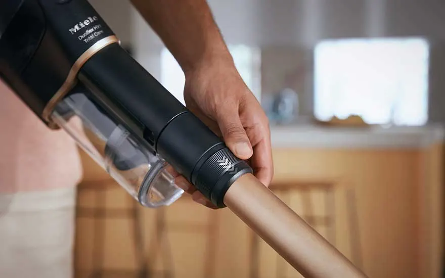 Een hand houdt een zwarte Miele stofzuiger met een doorzichtige stofbak vast. De stofzuiger heeft een gouden buis.