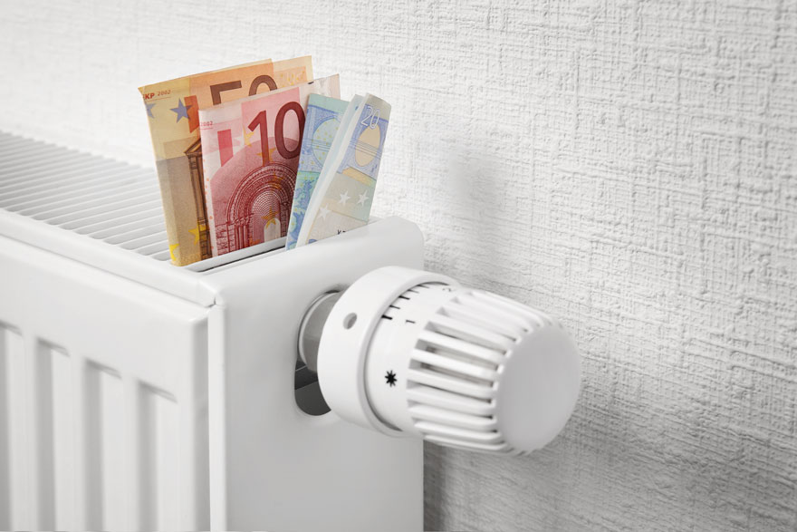 Een witte radiator met een thermostaatknop staat tegen een witte, getextureerde muur. Bovenop de radiator zitten verschillende eurobiljetten.