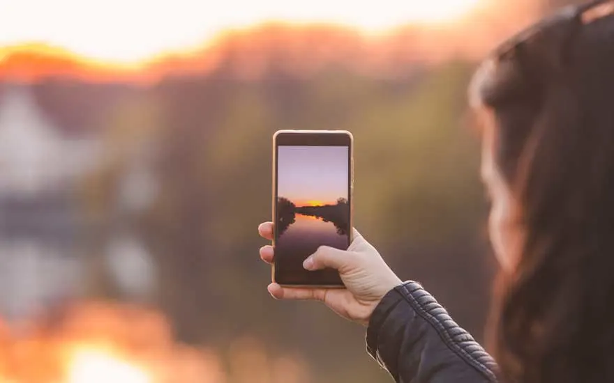 Une femme photographie un joli coucher de soleil sur une rivière avec son smartphone, devant un paysage flou et chaleureux.