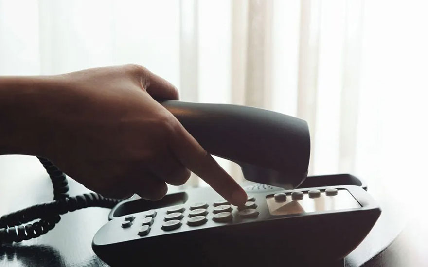 Een donkergrijze telefoon op een tafel, met een hand die een knop indrukt. Er is een witte gordijn op de achtergrond.