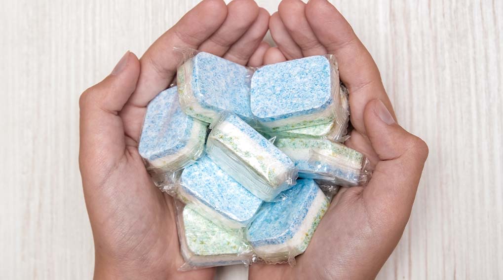 Twee handen houden een stapel afwasmachinetabletten, elk verpakt in doorzichtig plastic, tegen een houten achtergrond.