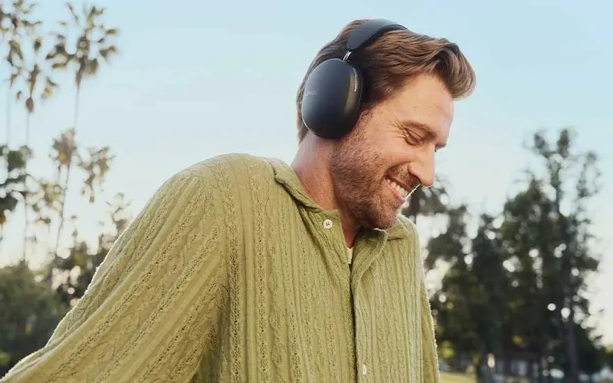 Un homme porte un casque audio noir et une chemise vert clair avec des arbres en arrière-plan.