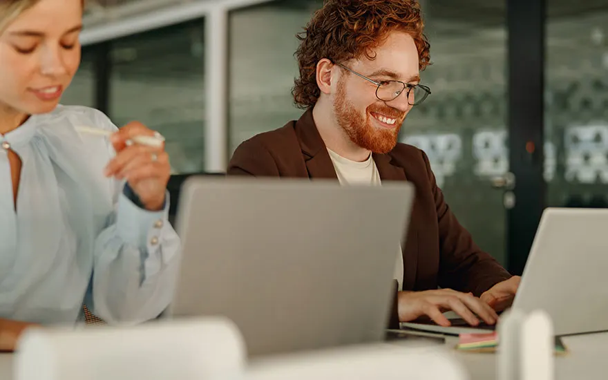 Twee mensen werken aan laptops op een bureau. De achtergrond is een kantoor.