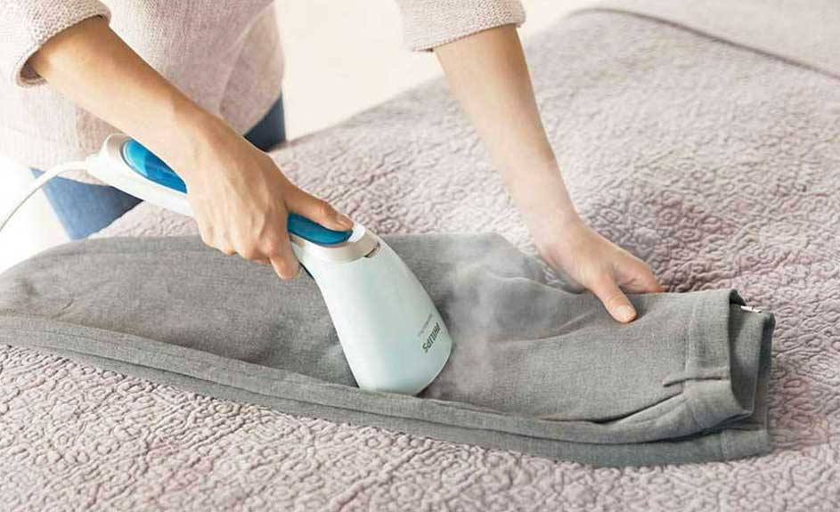 Een blauw-wit stoomstrijkijzer strijkt een grijze broek op een bed met een beigekleurige sprei.