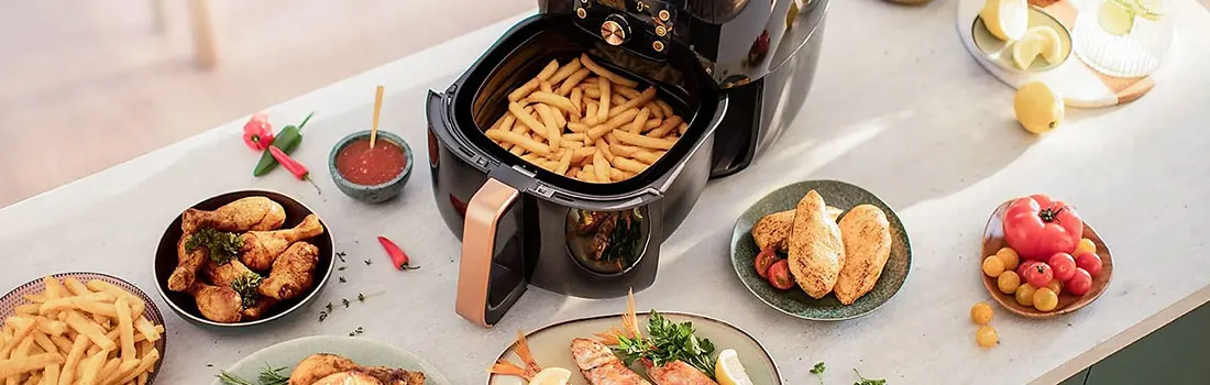 An air fryer filled with fries sits on a table with plates of chicken, fish, tomatoes, and a bowl of fries.