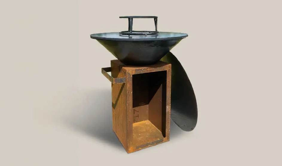 Een metalen vuurschaal of barbecue staat op een roestkleurige sokkel; ernaast leunt een ronde metalen plaat.