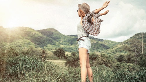 Een veld met groen gras en bergen. Er staat een persoon met open armen en een hoed, gekleed in een grijs hemd en blauwe short.