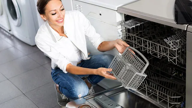 Une femme en chemise blanche et jean bleu s'accroupit devant un lave-vaisselle ouvert pour inspecter l'intérieur. Elle vérifie le panier tandis qu'elle est agenouillée devant l'appareil. La cuisine a un design moderne avec carrelage gris.