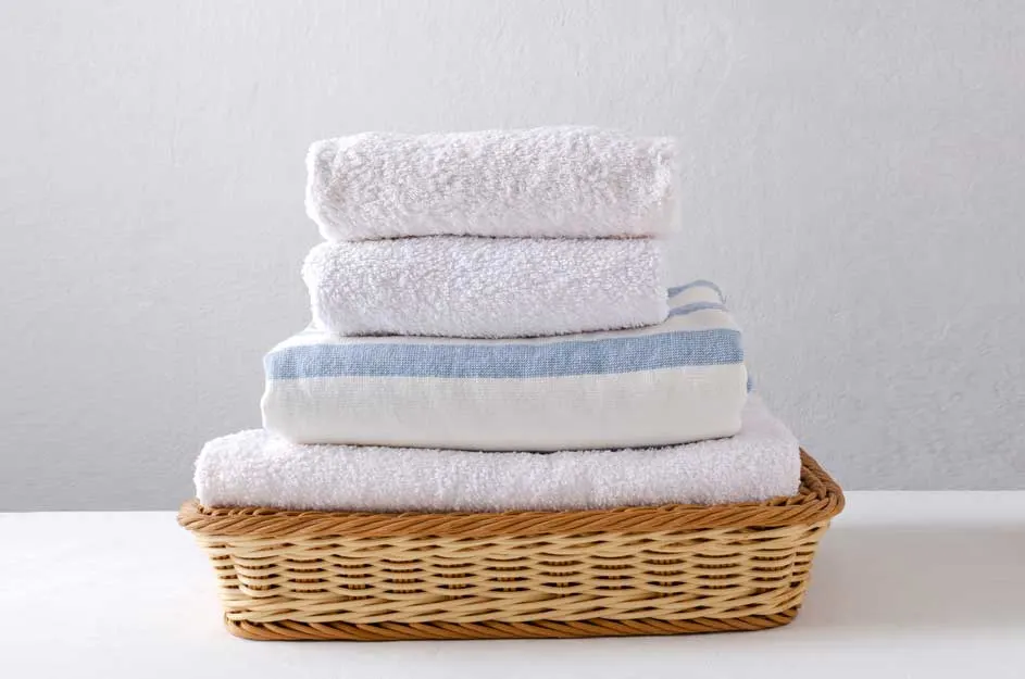 Une pile de serviettes blanches, dont une avec des rayures bleues, est posée dans un panier en osier. Le fond est blanc.
