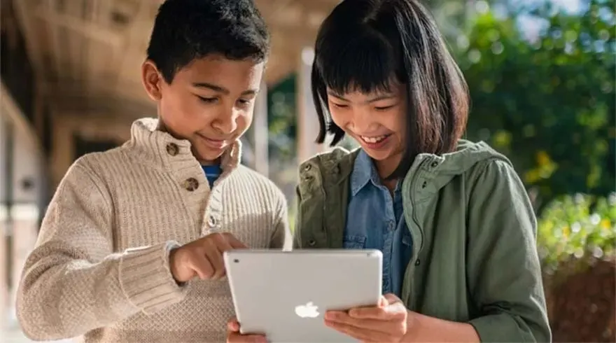 A silver tablet is held by two figures, one points to the screen. The background shows greenery and building detail.