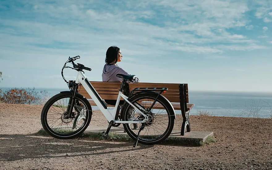 Een witte fiets staat naast een houten bank op een zandstrand, met uitzicht op de blauwe zee en lucht.