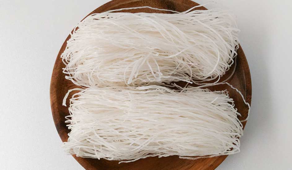 Deux portions de nouilles de riz blanches sur une assiette en bois brun, sur un fond blanc.