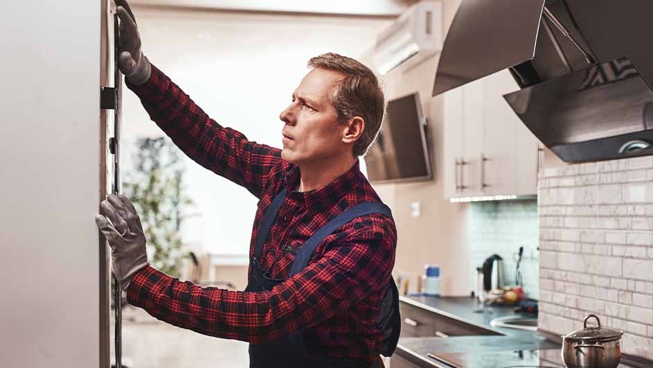 A person in workwear adjusts a white cabinet in a kitchen setting. Background shows countertops and appliances.