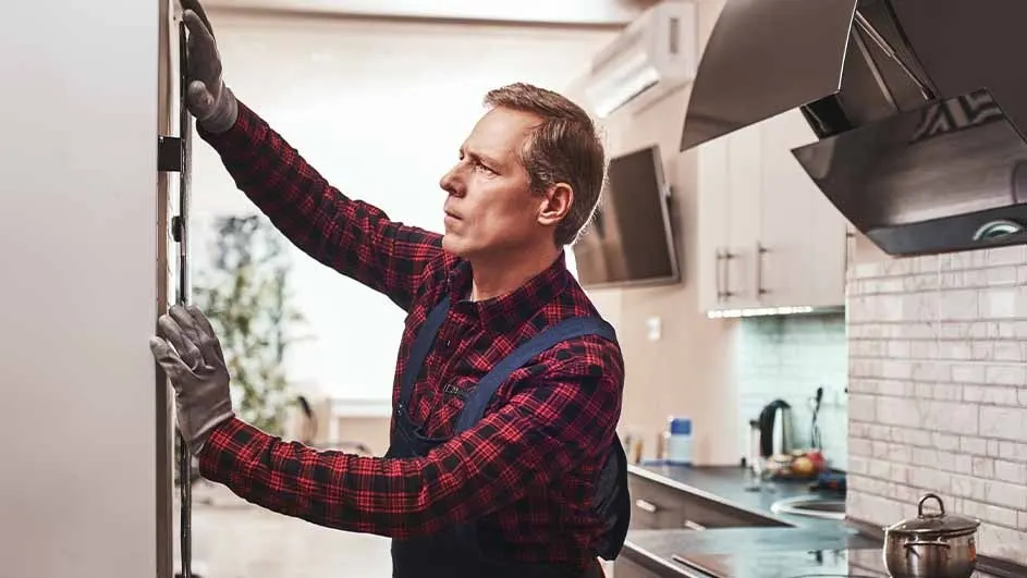 A person in workwear adjusts a white cabinet in a kitchen setting. Background shows countertops and appliances.