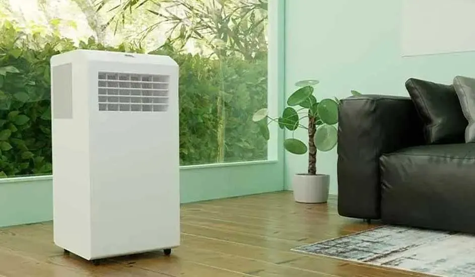 Een witte mobiele airco op wieltjes staat in een lichte kamer met houten vloer, naast een zwarte bank en een plant bij het raam.