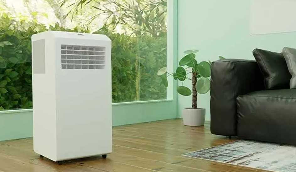 MMS World A white mobile air conditioner on wheels is positioned in a bright room with a wooden floor, next to a black sofa and a plant by the window.