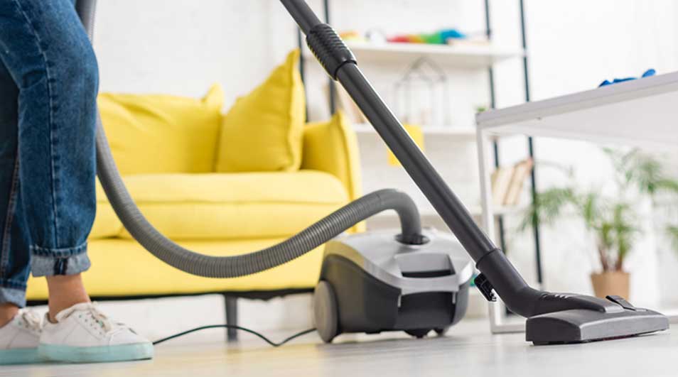 Aspirateur gris et noir sur sol clair, avec canapé jaune et étagères blanches en arrière-plan.