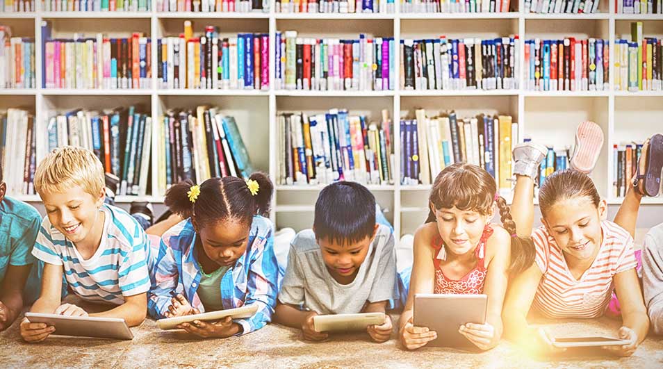 Vijf kinderen liggen op de grond met tablets, voor een witte boekenkast vol boeken.