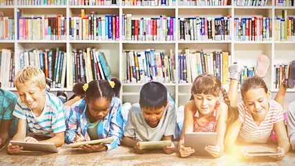 Vijf kinderen liggen op de grond met tablets, voor een witte boekenkast vol boeken.