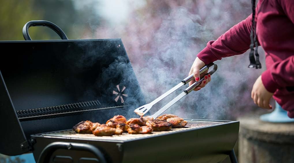 A black grill with the lid open shows food cooking on the grates. Tongs are turning the food with smoke rising.