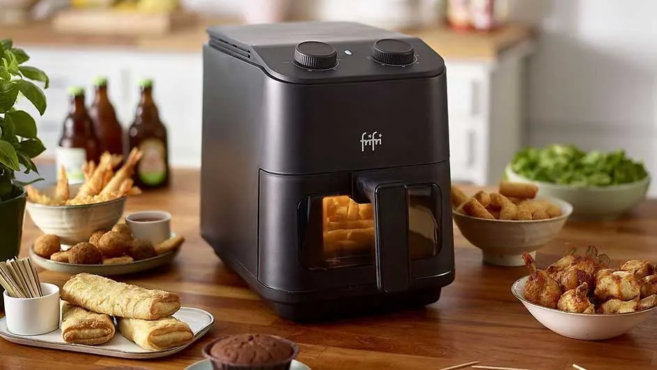 Zwarte friteuse op een houten tafel, omringd door gefrituurde hapjes, bierflessen en een groene plant.