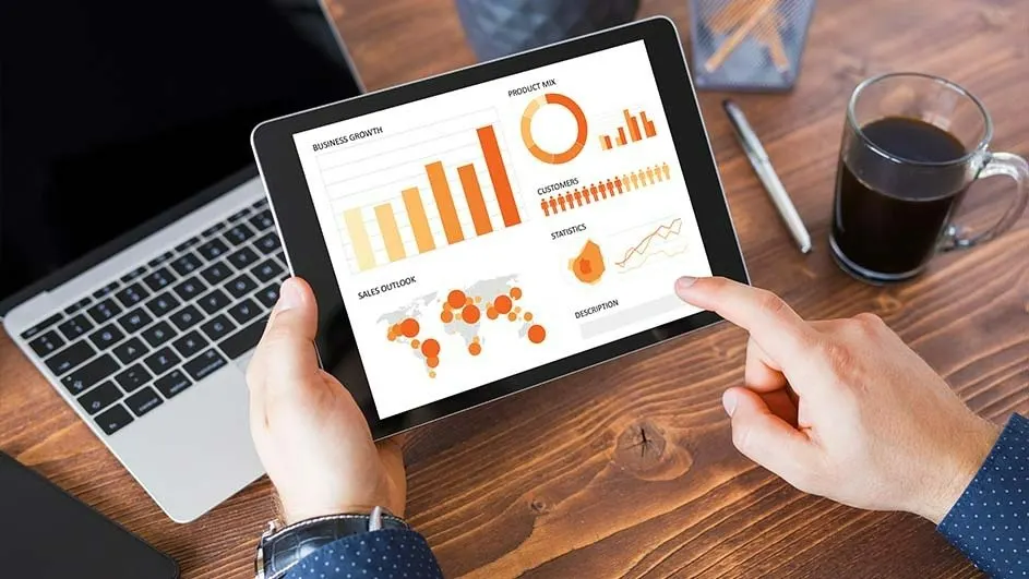 Een tablet toont grafieken over zakelijke groei naast een laptop, koffie en een pen op een houten bureau.