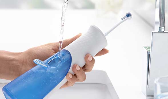Quelqu’un remplit un hydropulseur blanc et bleu au robinet; l’eau coule dans le réservoir, embout levé, salle de bain lumineuse.