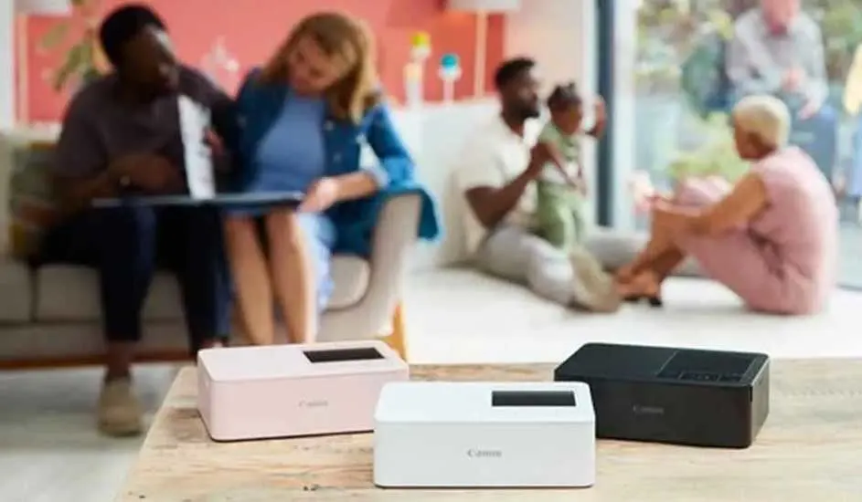 Drie Canon-printers in wit, roze en zwart op een houten tafel. Op de achtergrond zit een familie in een woonkamer.