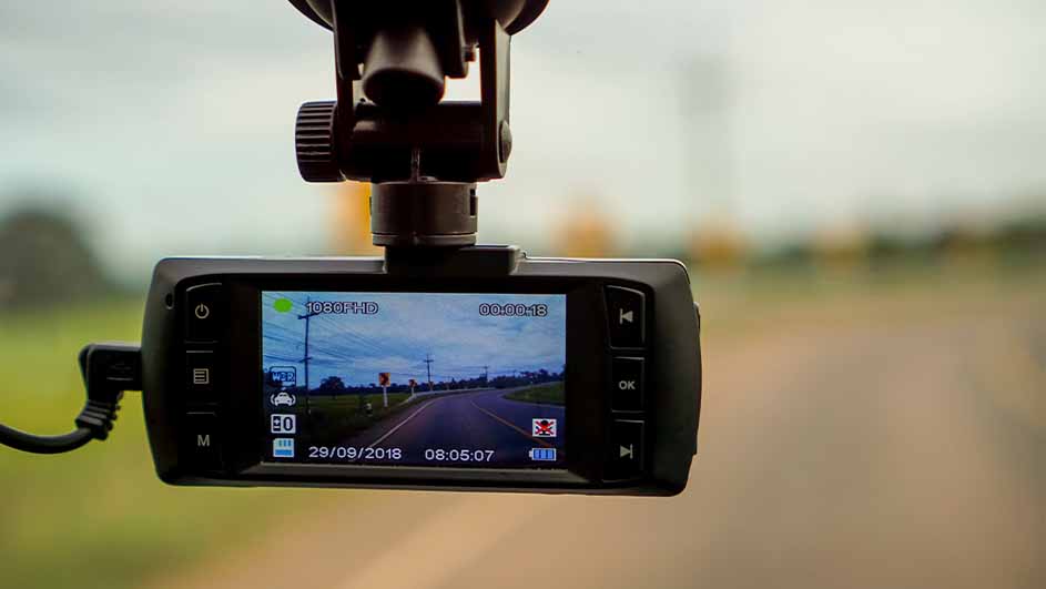 Zwarte dashcam bevestigd aan een autoruit, met een scherm dat een weg laat zien. Datum en tijd zijn zichtbaar.