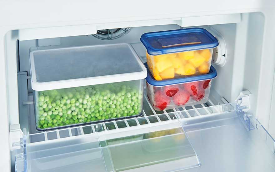 Dans un frigo blanc, trois récipients transparents remplis de pois, mangues et fraises, avec des couvercles blancs et bleus.