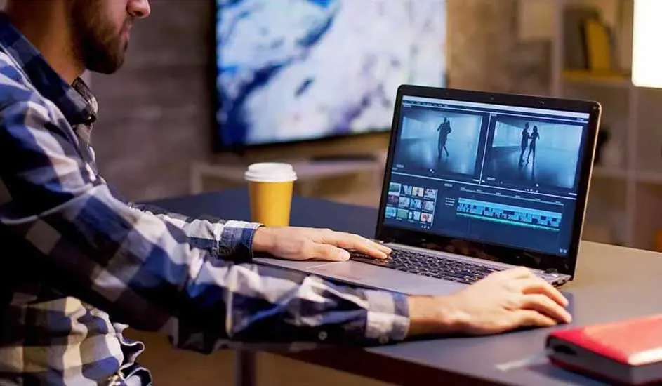Man gebruikt laptop voor videobewerking. Beker en rode harde schijf op bureau. TV aan de muur.