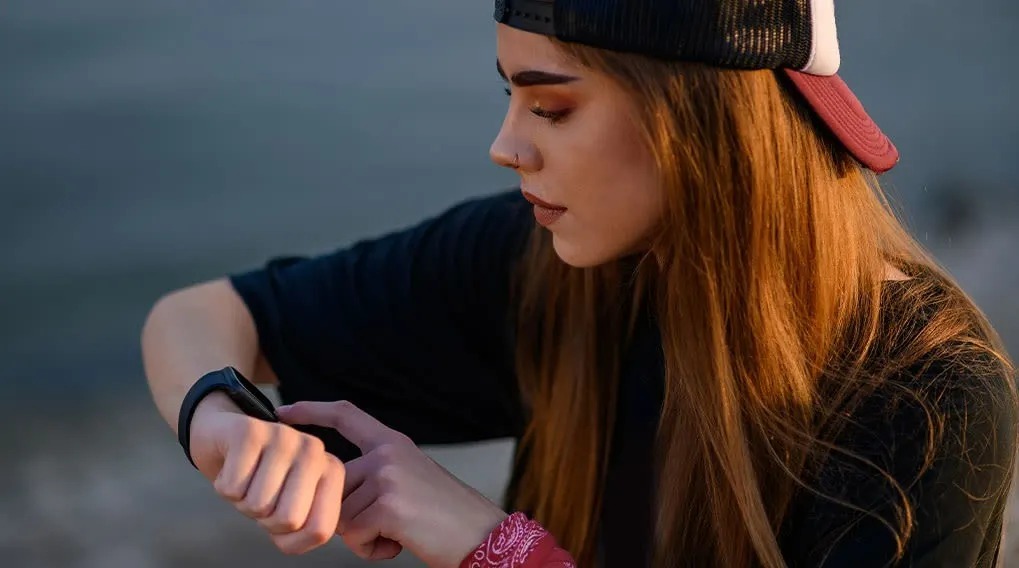 A person with long red hair, a cap, and a black shirt is operating a black smartwatch on their wrist outdoors, near a body of water.