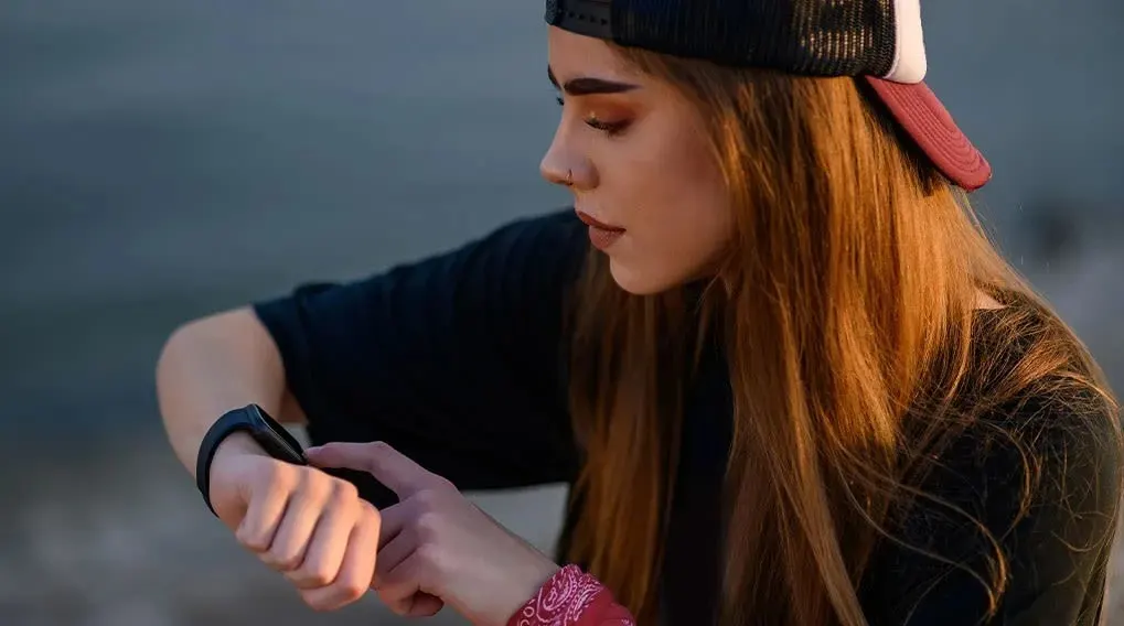 A person with long red hair, a cap, and a black shirt is operating a black smartwatch on their wrist outdoors, near a body of water.